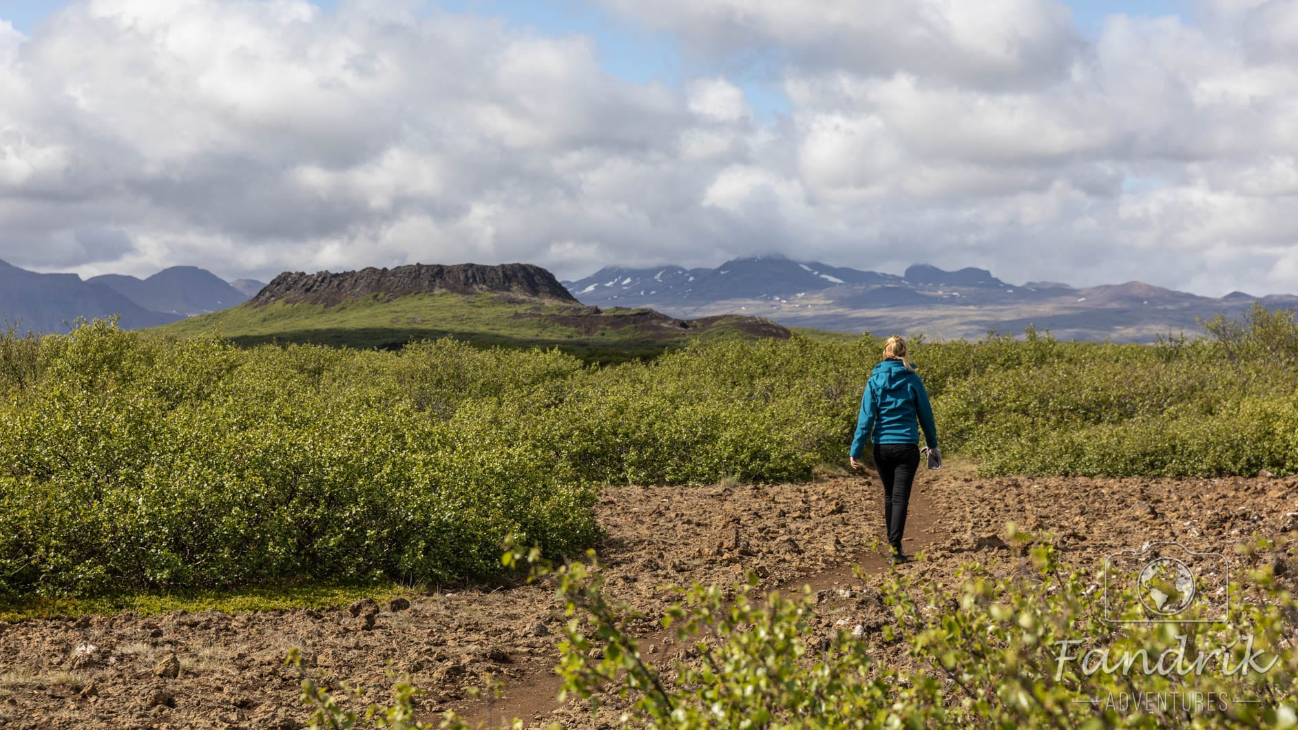 Eine Frau wandert auf einem steinigen Pfad zum Eldborg Krater, welcher im Hintergrund aufragt.